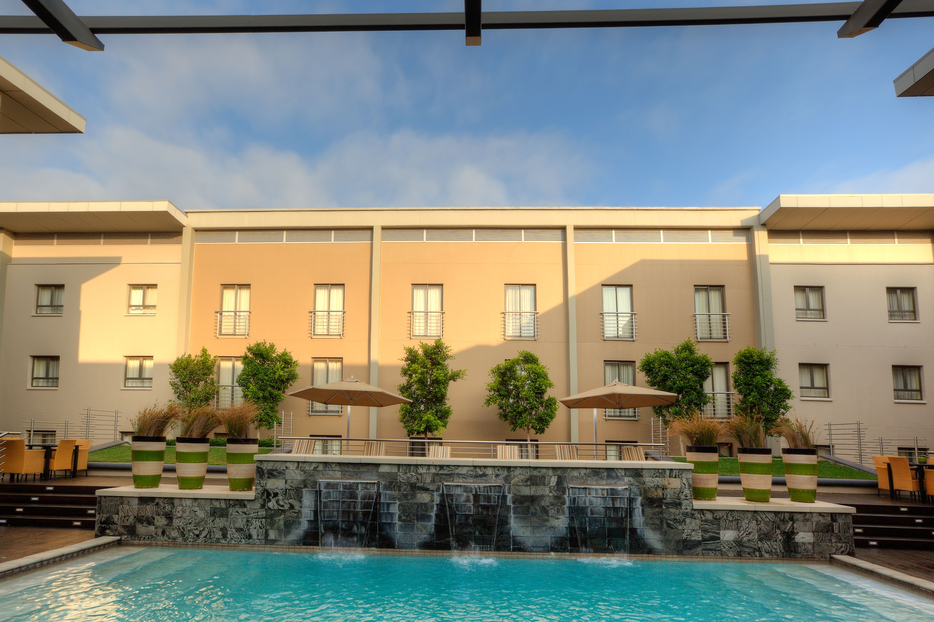 A hotel pool area with a waterfall feature, outdoor seating, and a view of the building facade.