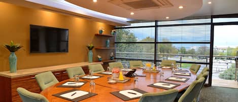 Un salón de conferencias con una gran mesa ovalada, múltiples sillas, un televisor de pantalla plana montado en la pared y vistas al exterior a través de amplias ventanas.