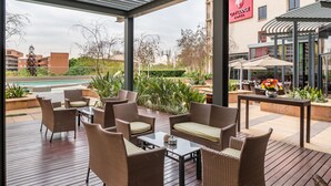 A covered outdoor seating area with wicker furniture and a view of a pool and buildings in the background. - City Lodge Hotel Hatfield (Pretoria)