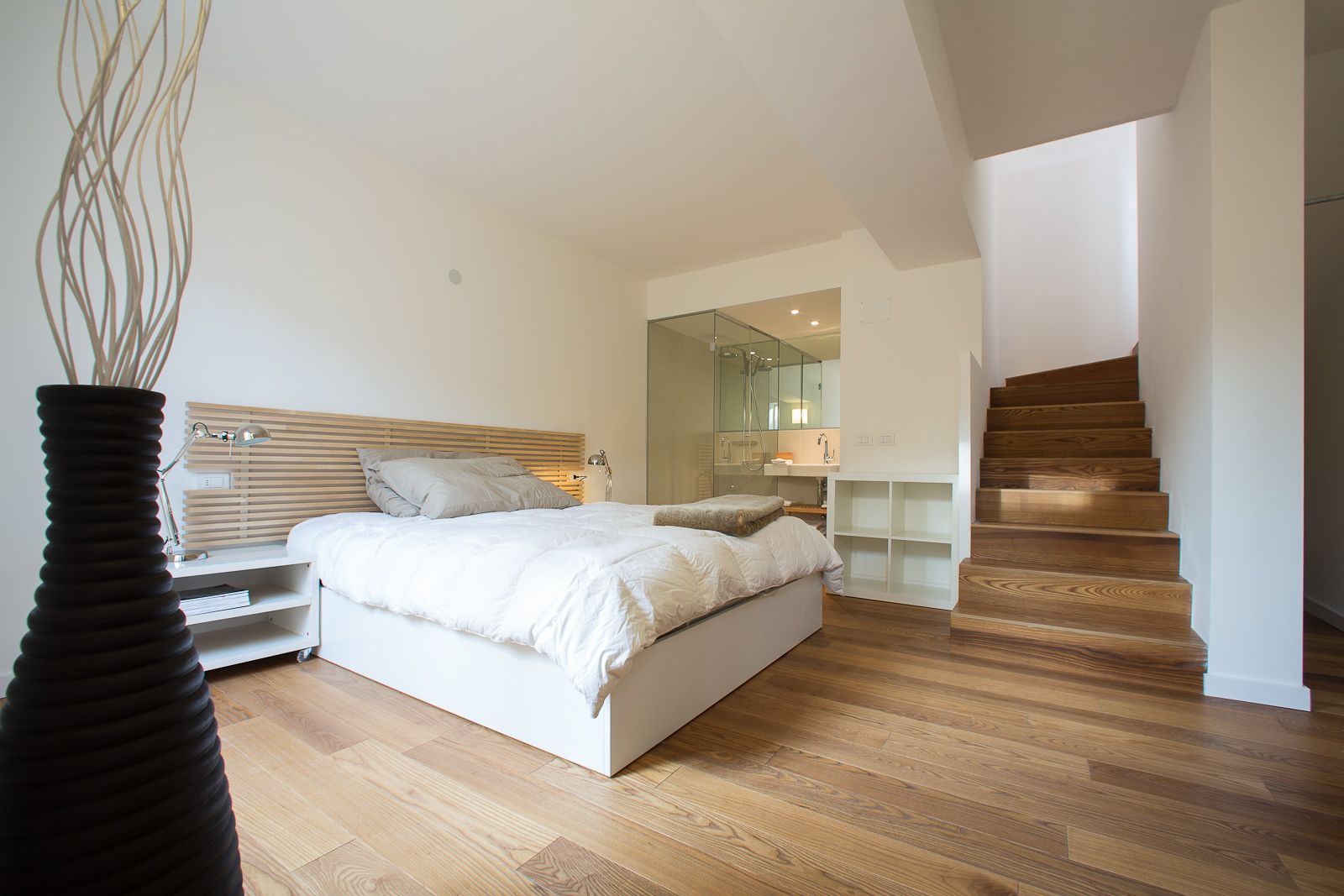 A modern bedroom with a wooden floor, a bed with white bedding, a nightstand, a bedside table, a black vase, and a staircase leading to an upper level.