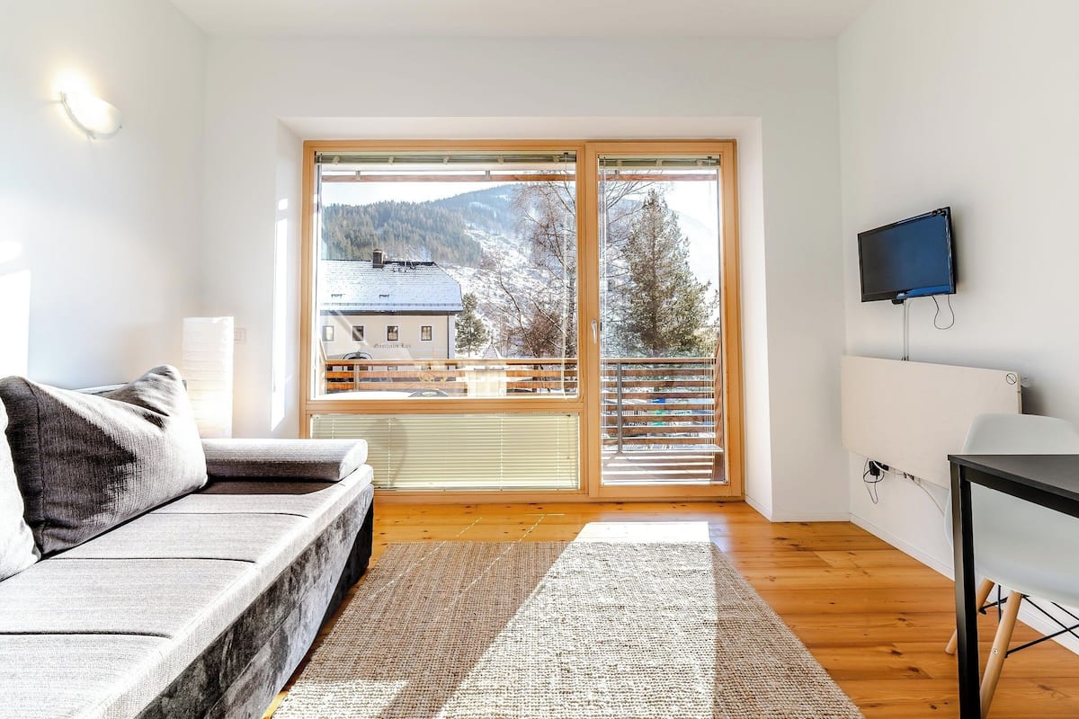 A modern living room with a sofa, a flat-screen TV mounted on the wall, and a large window offering a view of a snowy landscape and a building.