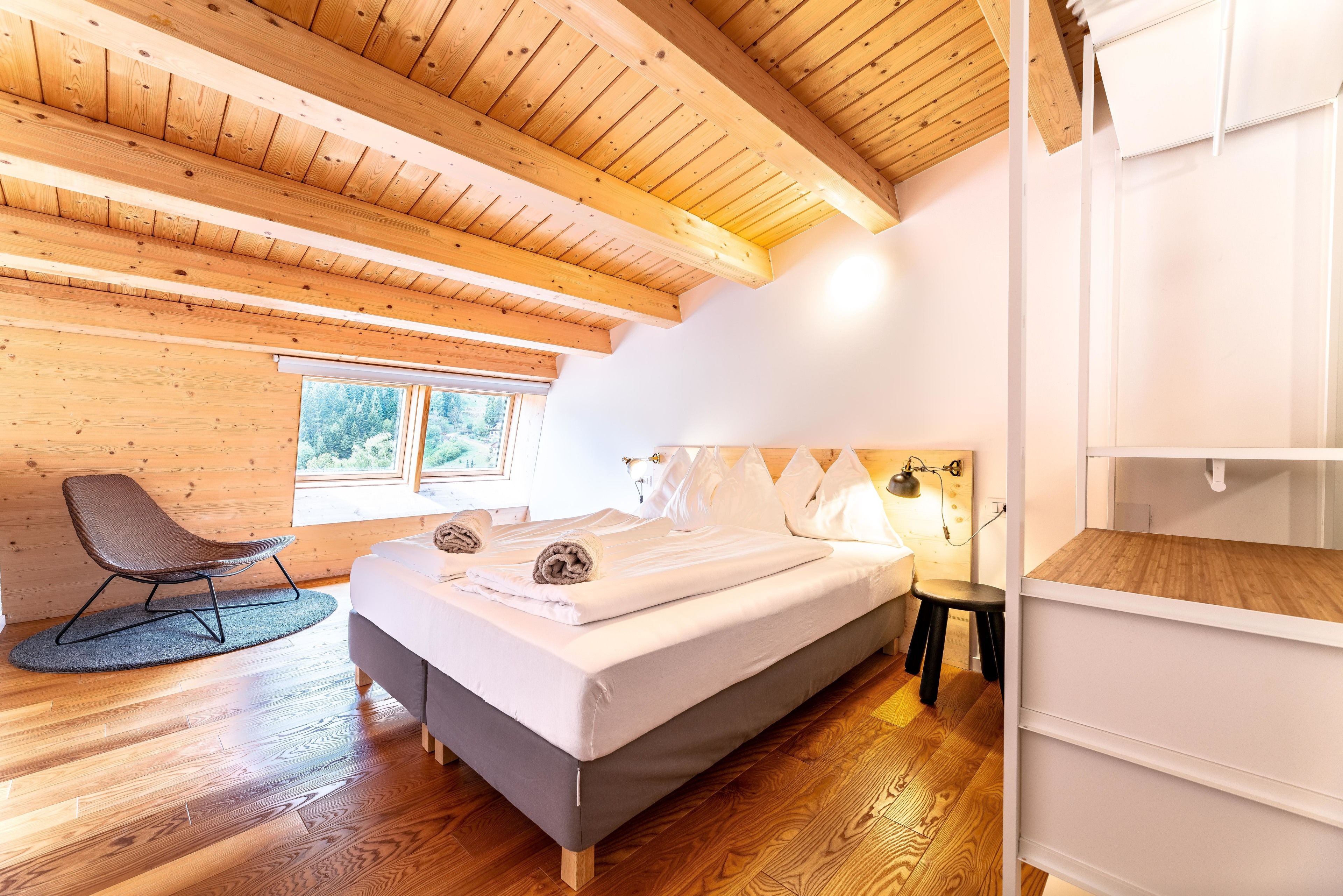 A bedroom with a wooden ceiling, a bed with pillows, a chair, and a bedside table.