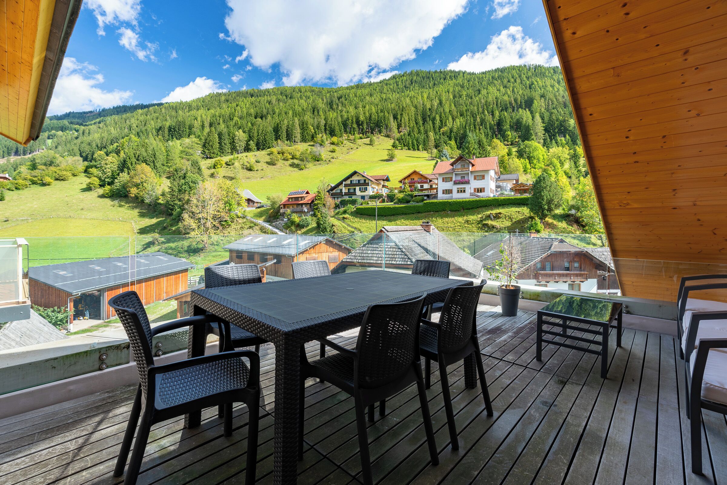 Eine Holzdar Terrasse mit Tisch und Stühlen, mit Blick auf ein grünes Tal mit Häusern und einem bewaldeten Hügel.
