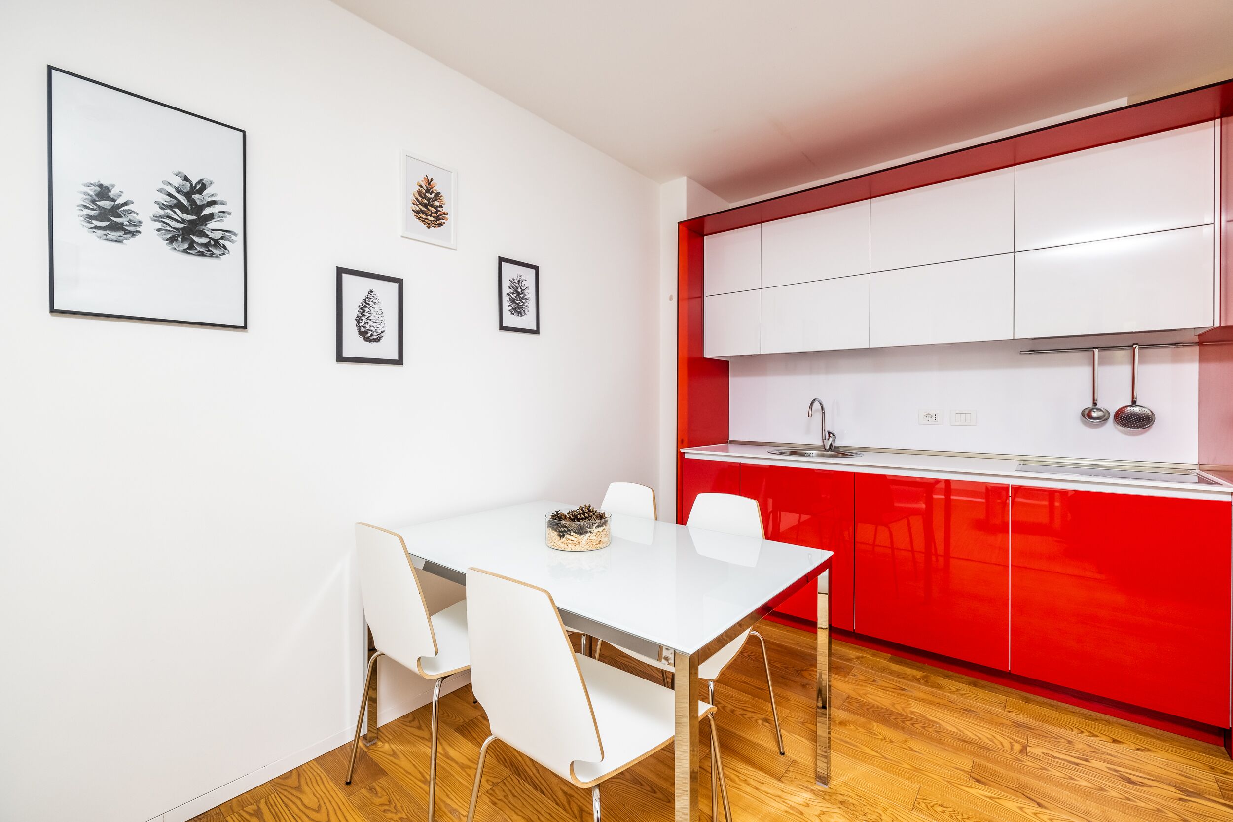 A modern kitchen with white cabinets and a red accent wall, a dining area with a white table and chairs, and framed artwork on the wall.