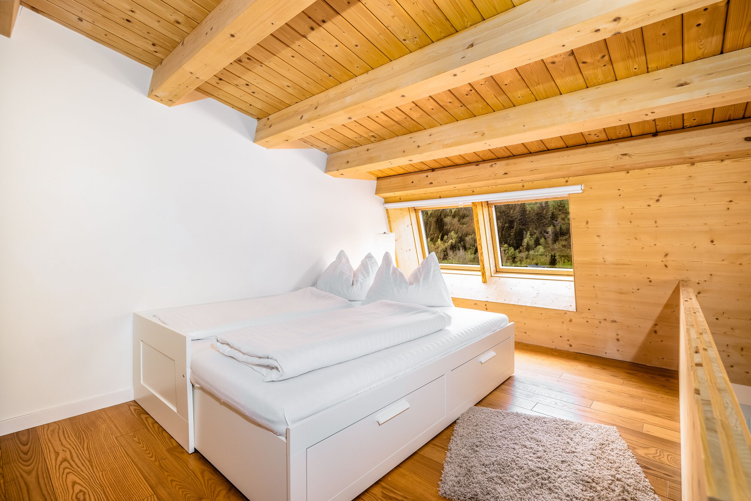 A bedroom with a wooden ceiling, a white bed, and a window with a view of trees.