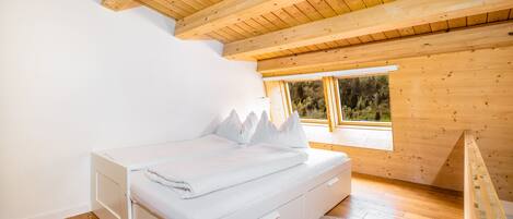 A bedroom with a wooden ceiling, a white bed, and a window with a view of trees.