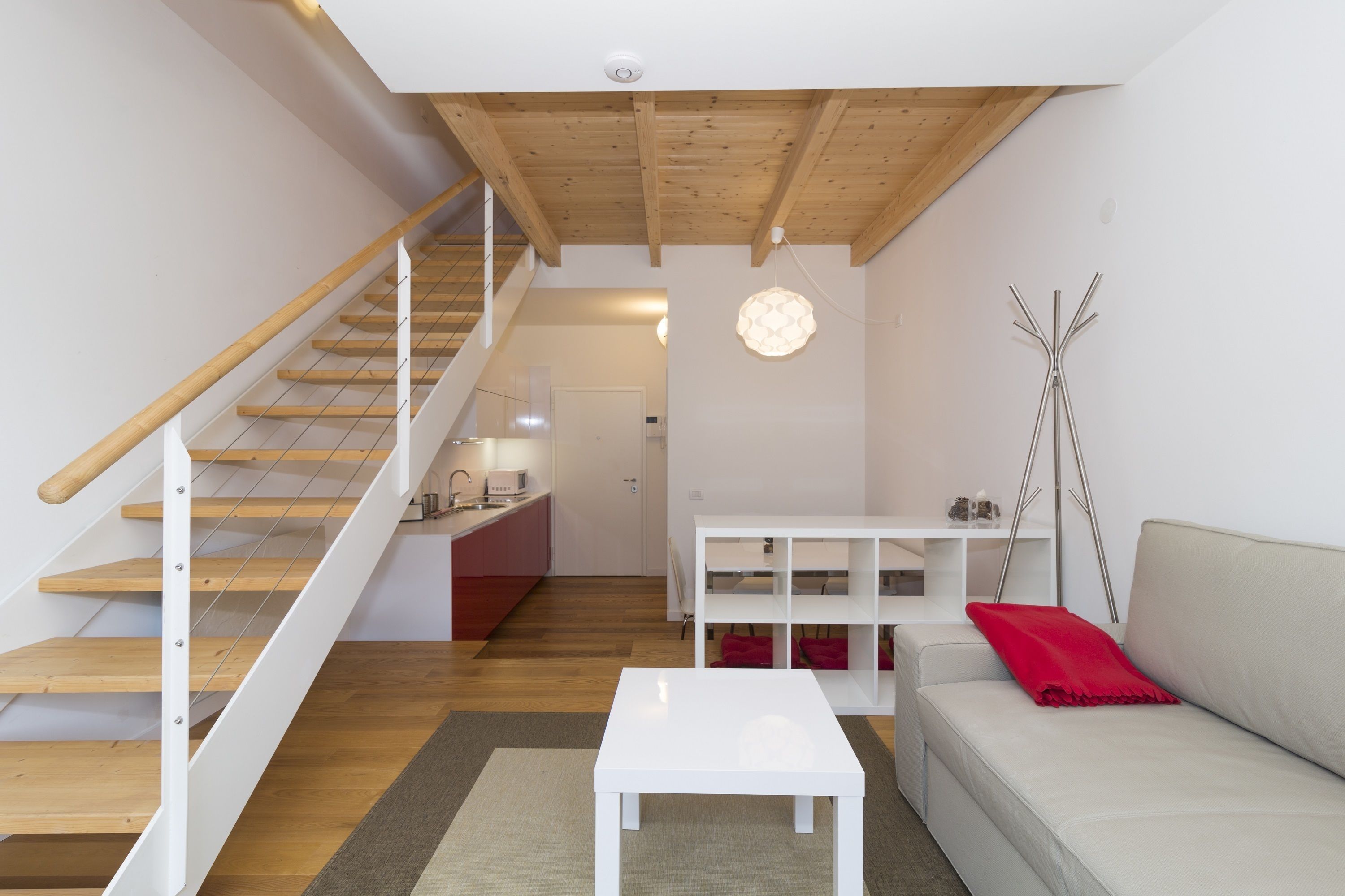 A modern living room with a staircase, a white sofa, a small table, and a kitchen area in the background.