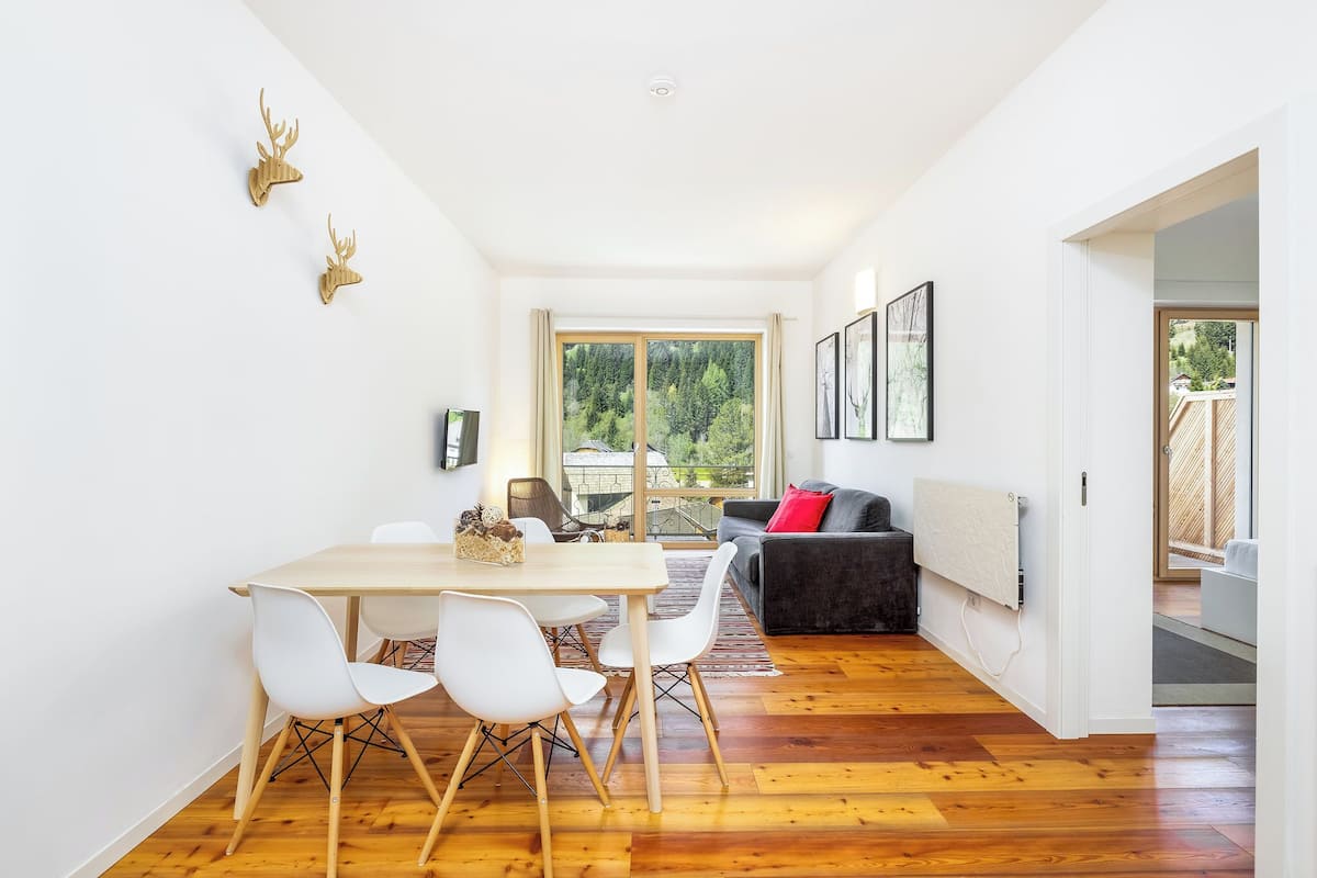 A modern dining area with a wooden table and white chairs, a black sofa, and a large window with a view of trees.