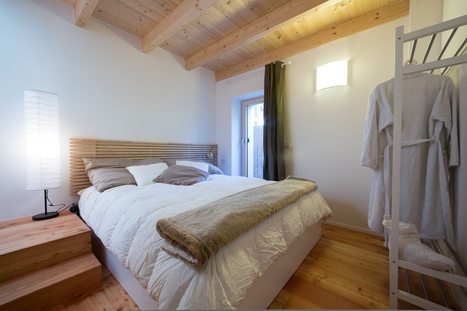Un dormitorio con cama de cabecera de madera, lámpara de noche, ventana con cortinas y un perchero metálico con toallas.