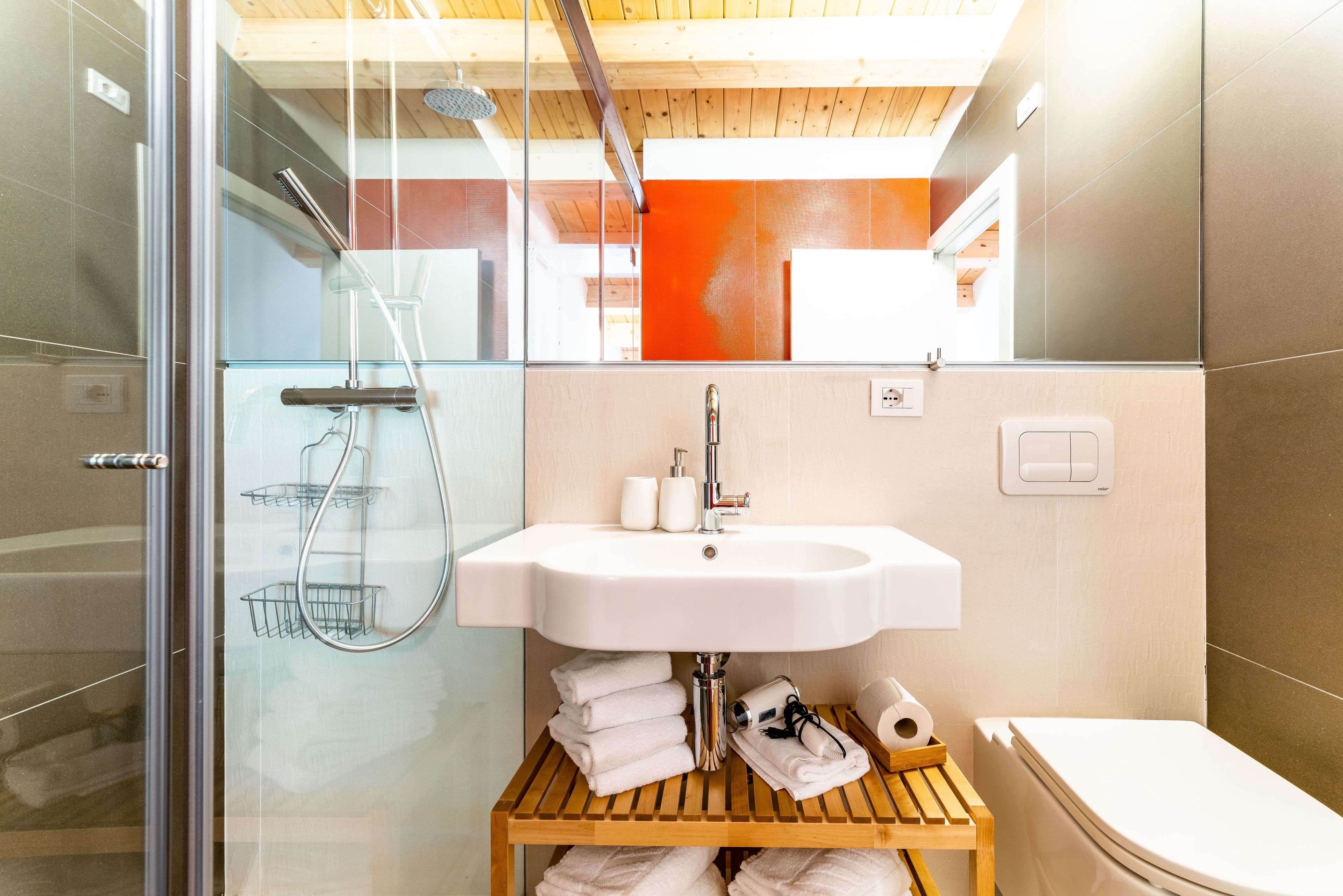 A modern bathroom with a glass shower enclosure, a white sink, and a wooden shelf with towels.