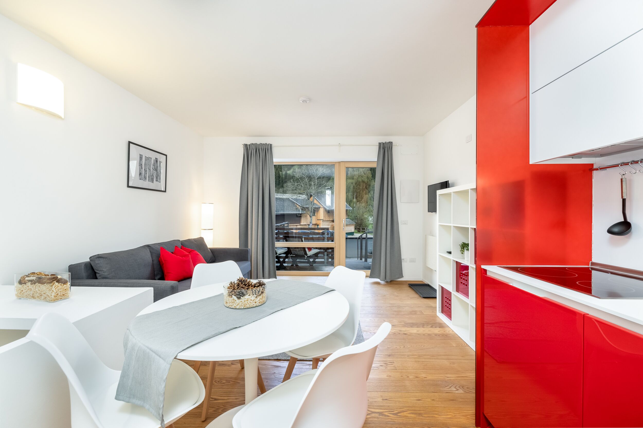 A modern living room with a dining area, a kitchen with red cabinets, and a view through a large window.