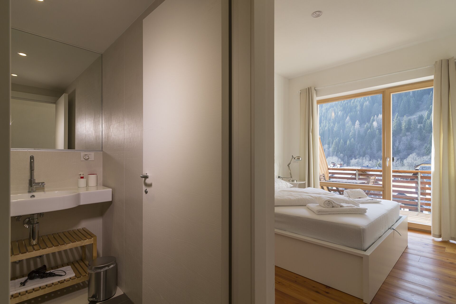 A bedroom with a bed, a nightstand, a window with a view of trees, and a bathroom with a sink and mirror.