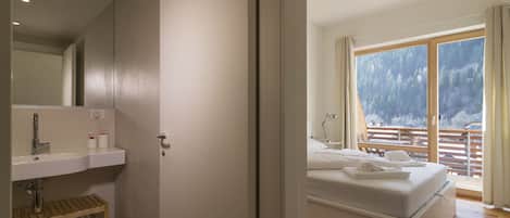 A bedroom with a bed, a nightstand, a window with a view of trees, and a bathroom with a sink and mirror.