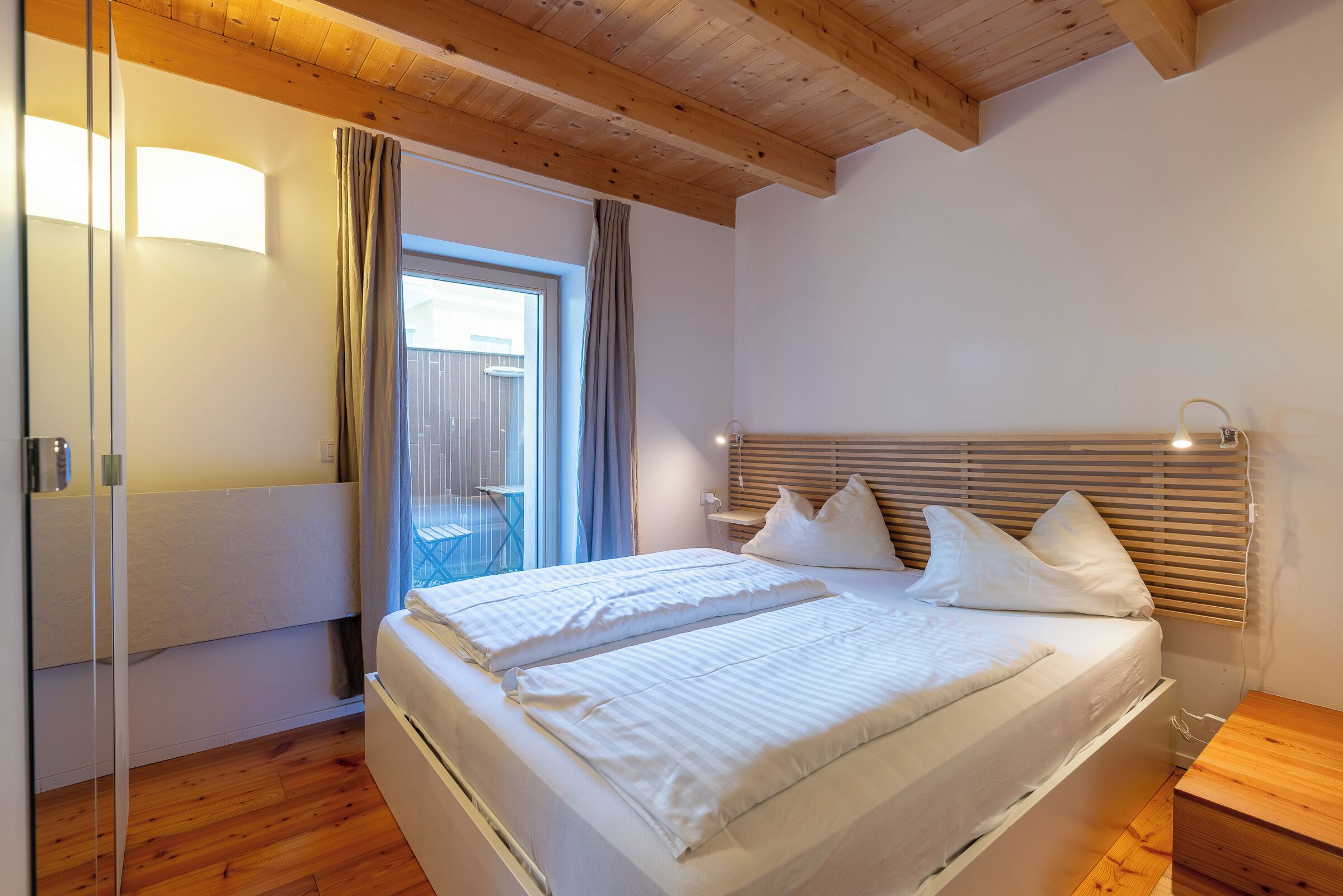Une chambre avec un plafond en bois, un lit avec une literie blanche, une table de chevet et une fenêtre avec des rideaux.