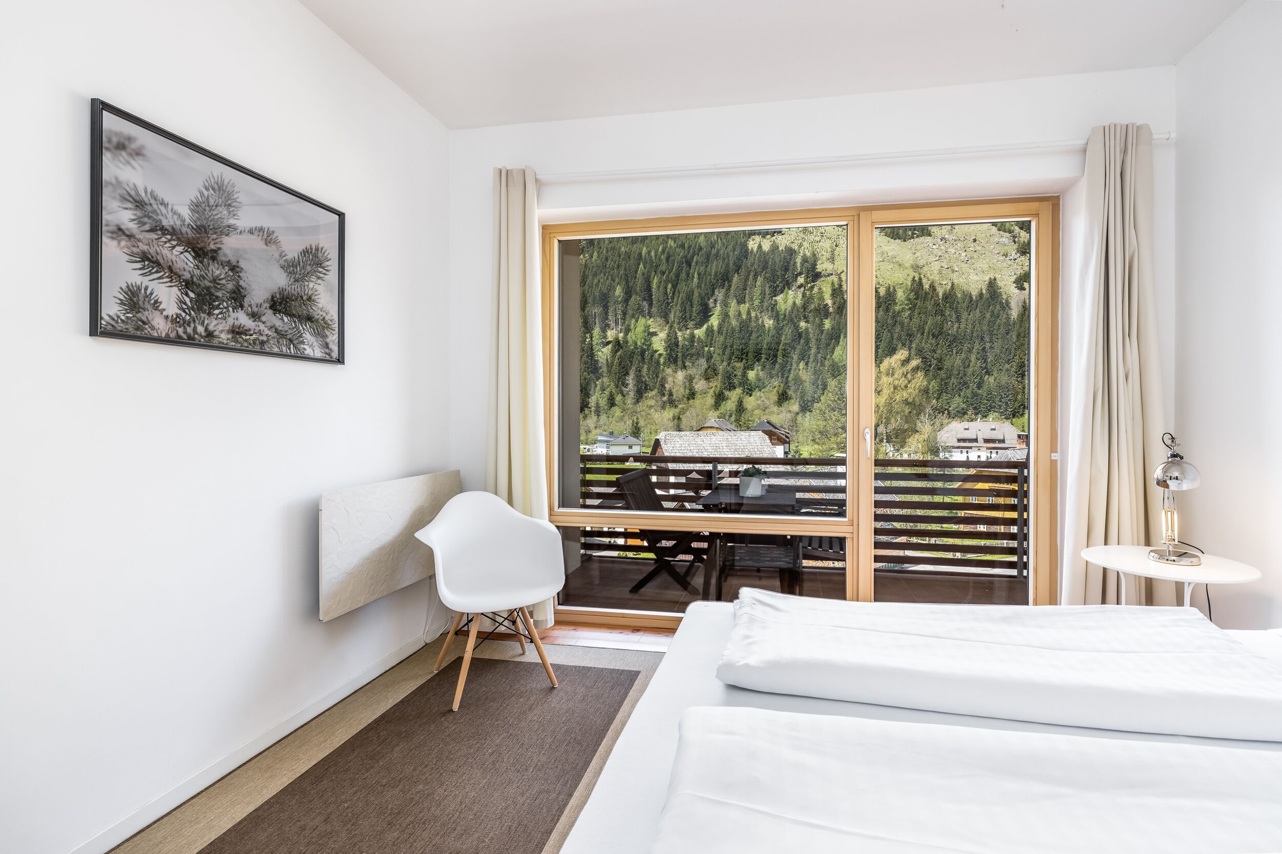 A hotel room with a bed, a desk, a chair, a window with a view of a forest and a mountain, and a painting on the wall.