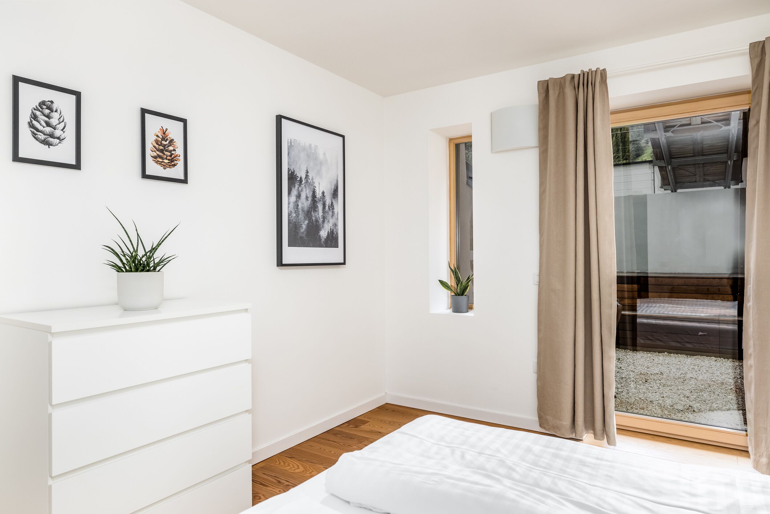 Une chambre à coucher avec un lit, une commode blanche, une fenêtre avec des rideaux et des tableaux encadrés accrochés au mur.