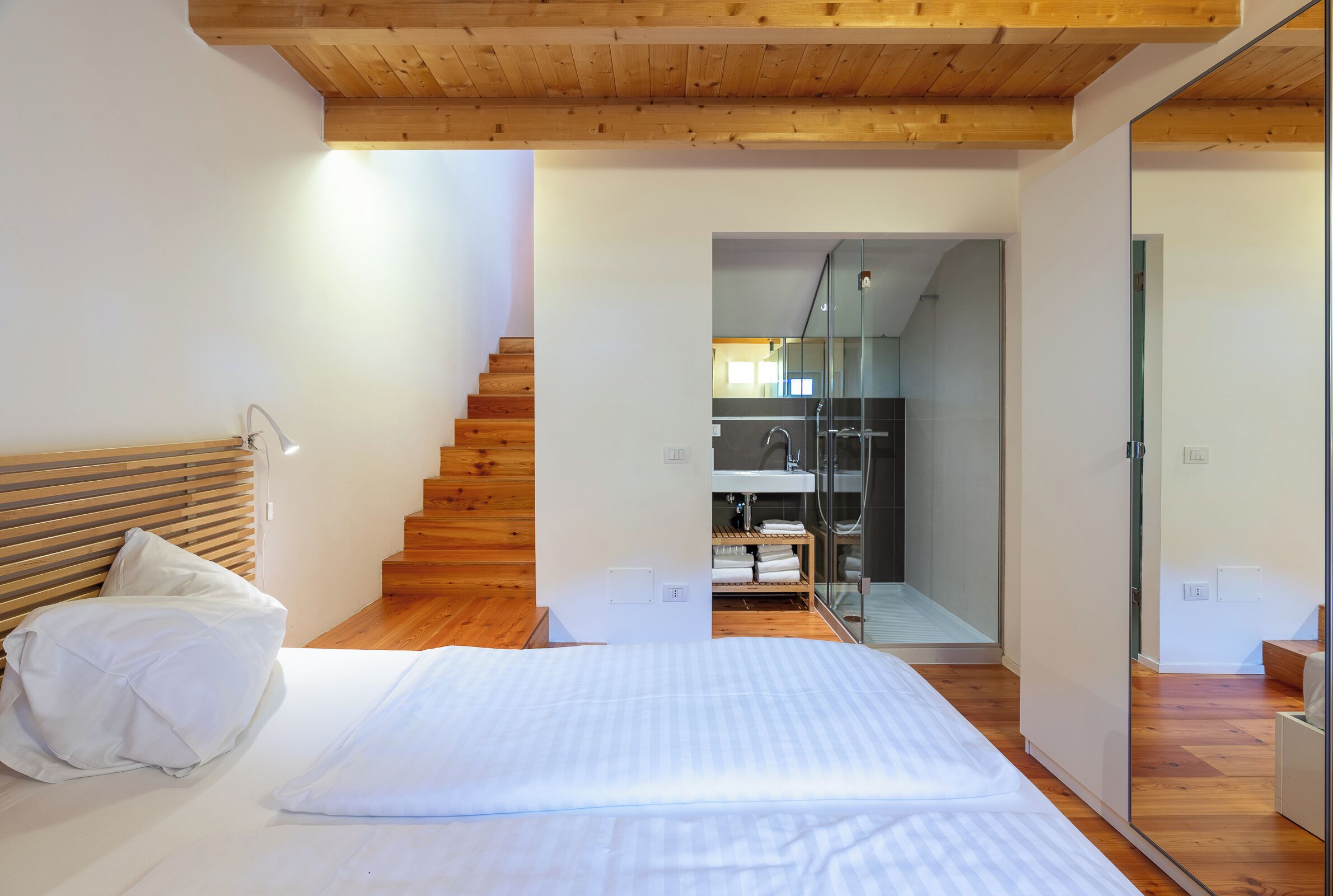 Un dormitorio con cabecera de madera, cama con ropa de cama blanca, escalera de madera y zona de ducha de vidrio.