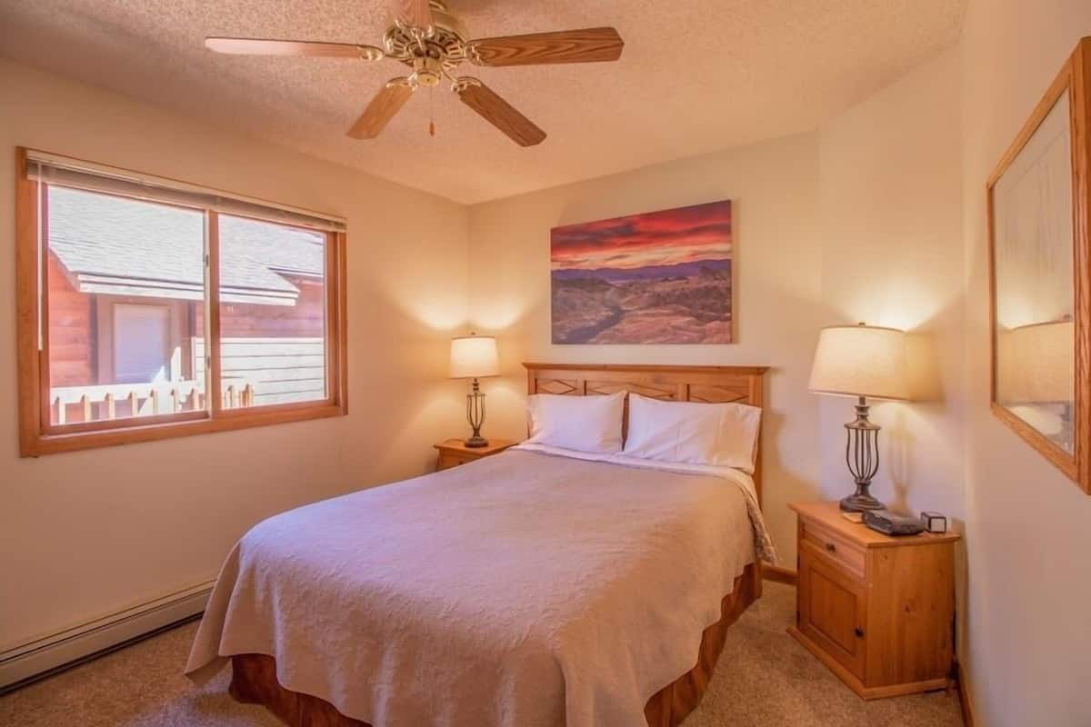 A bedroom with a bed, two bedside lamps, a ceiling fan, a window with blinds, and a painting on the wall.