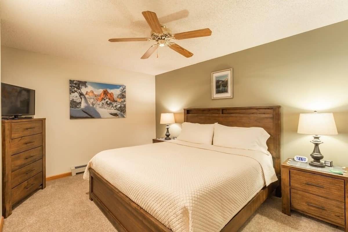 A bedroom with a wooden bed, two bedside tables with lamps, a ceiling fan, and two wall-mounted TVs.