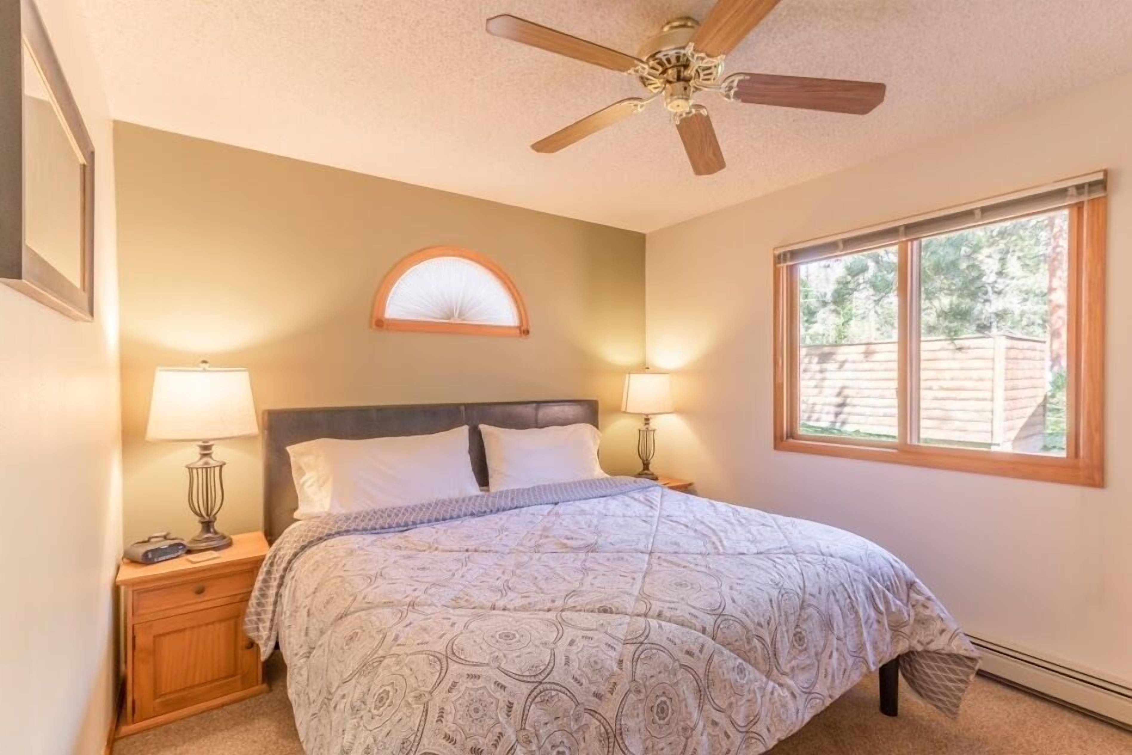 A bedroom with a bed, two bedside tables with lamps, a ceiling fan, a window with a view of the outside, and a wall-mounted mirror.