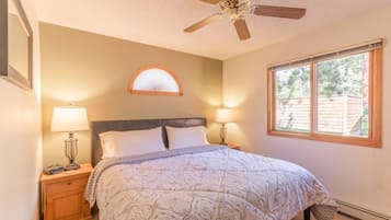 Un dormitorio con cama, dos mesitas de noche con lámparas, ventilador de techo, ventana con vistas al exterior y espejo adosado a la pared.