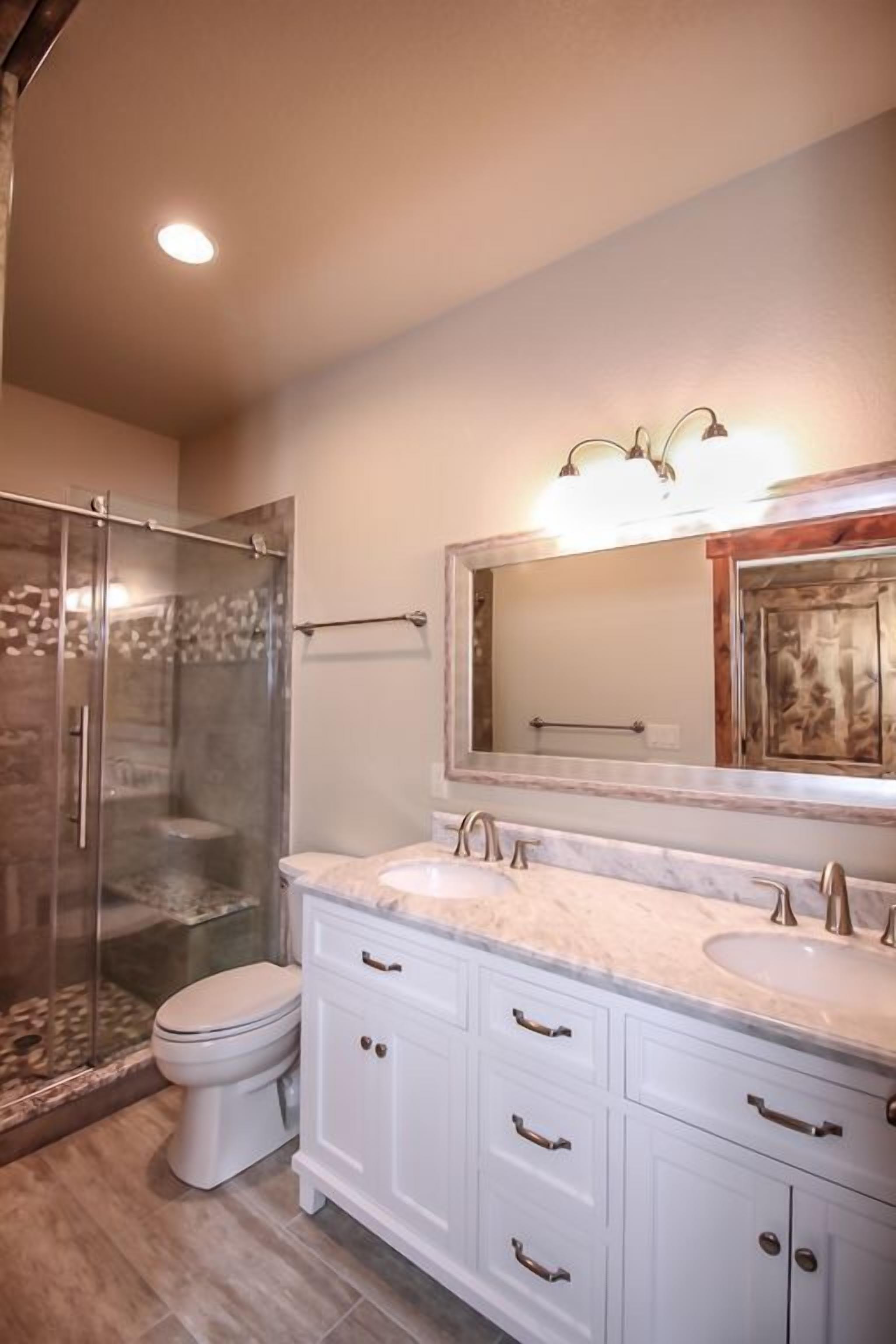A bathroom with a glass-enclosed shower, a white toilet, and a vanity with a large mirror and two sinks.