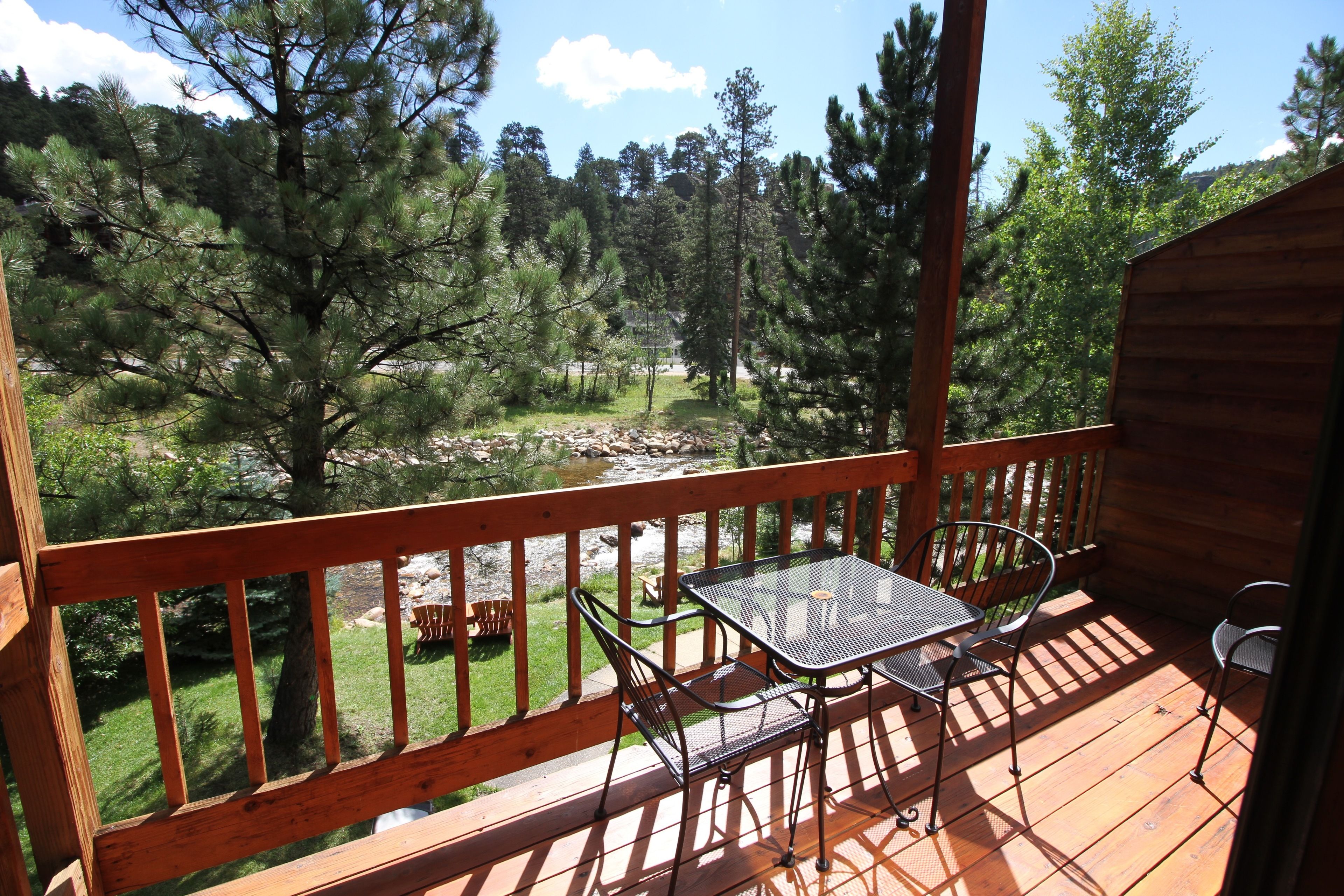 A wooden deck with a table and chairs, overlooking a natural landscape with trees and a stream.