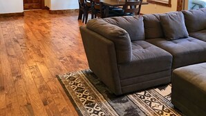 A spacious living room with a large gray sectional sofa, a dining area with a wooden table and chairs, and a large window with multiple panes. - Woodlands on Fall River (Estes Park)