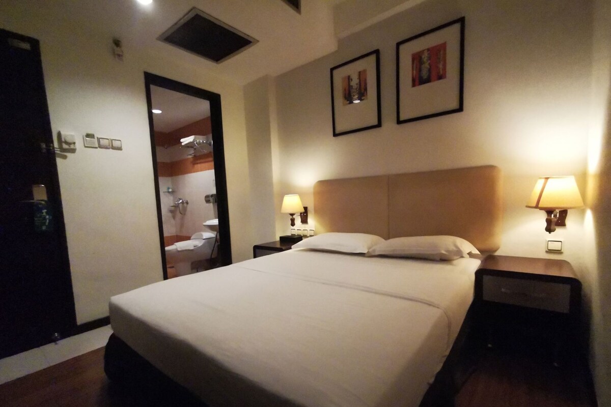 Una habitación de hotel con una cama, dos lámparas de noche, un cabecero, una puerta y un espejo.