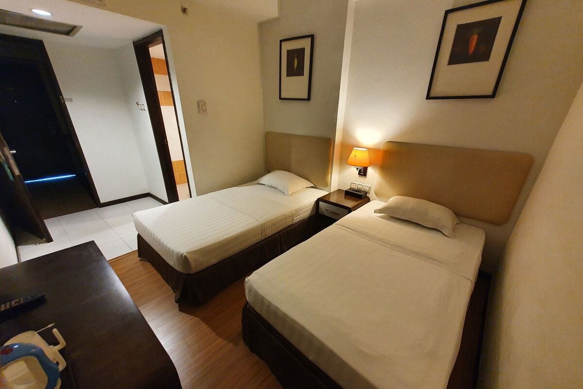 Una habitación de hotel con dos camas individuales, una mesita de noche con lámpara, una pequeña mesa con mando a distancia y una puerta que da a otra habitación.