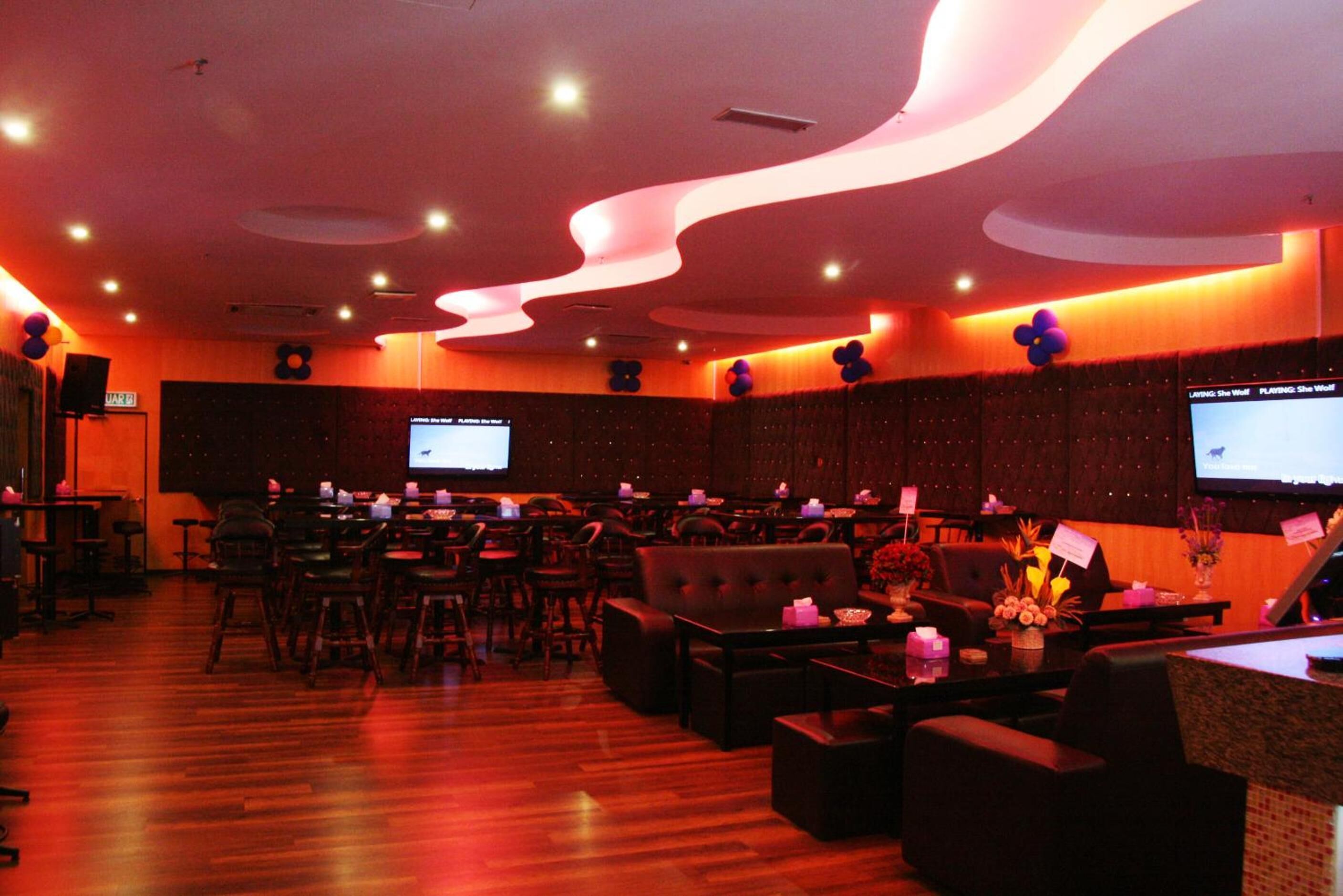 A spacious lounge area with multiple seating options, including sofas and chairs, arranged around a central table. There are several screens mounted on the walls, likely for presentations or information displays. The room features a wooden floor and a modern ceiling design with integrated lighting.
