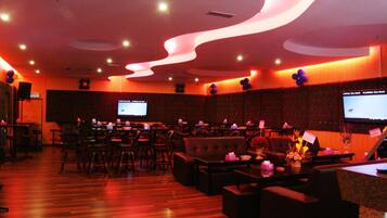 A spacious lounge area with multiple seating options, including sofas and chairs, arranged around a central table. There are several screens mounted on the walls, likely for presentations or information displays. The room features a wooden floor and a modern ceiling design with integrated lighting.