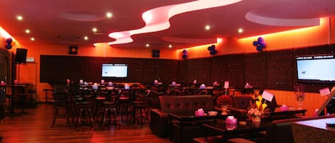 A spacious lounge area with multiple seating options, including sofas and chairs, arranged around a central table. There are several screens mounted on the walls, likely for presentations or information displays. The room features a wooden floor and a modern ceiling design with integrated lighting.