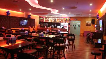Una zona de bar con mesas y sillas de madera, una pantalla de televisión y una barra con diferentes botellas y vasos.
