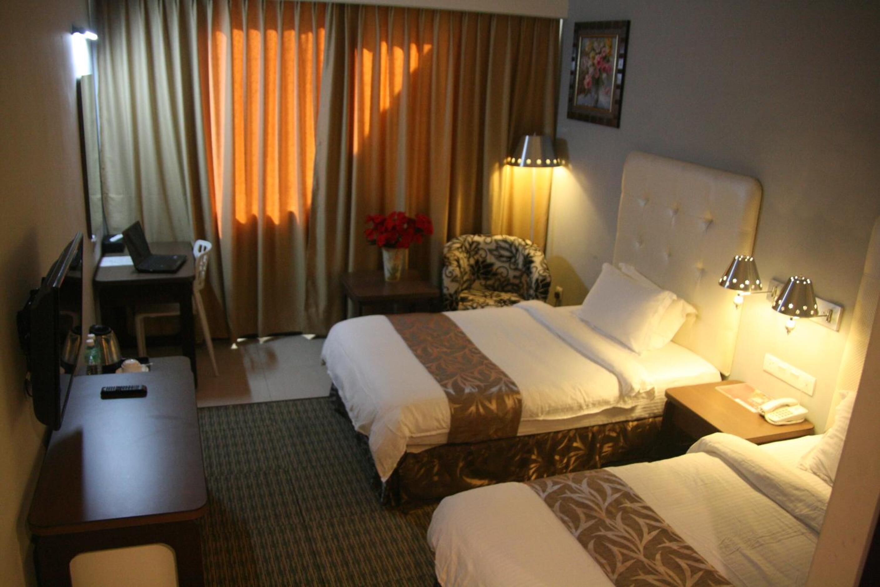 A hotel room with two beds, a desk, a chair, a lamp, and a vase with flowers.