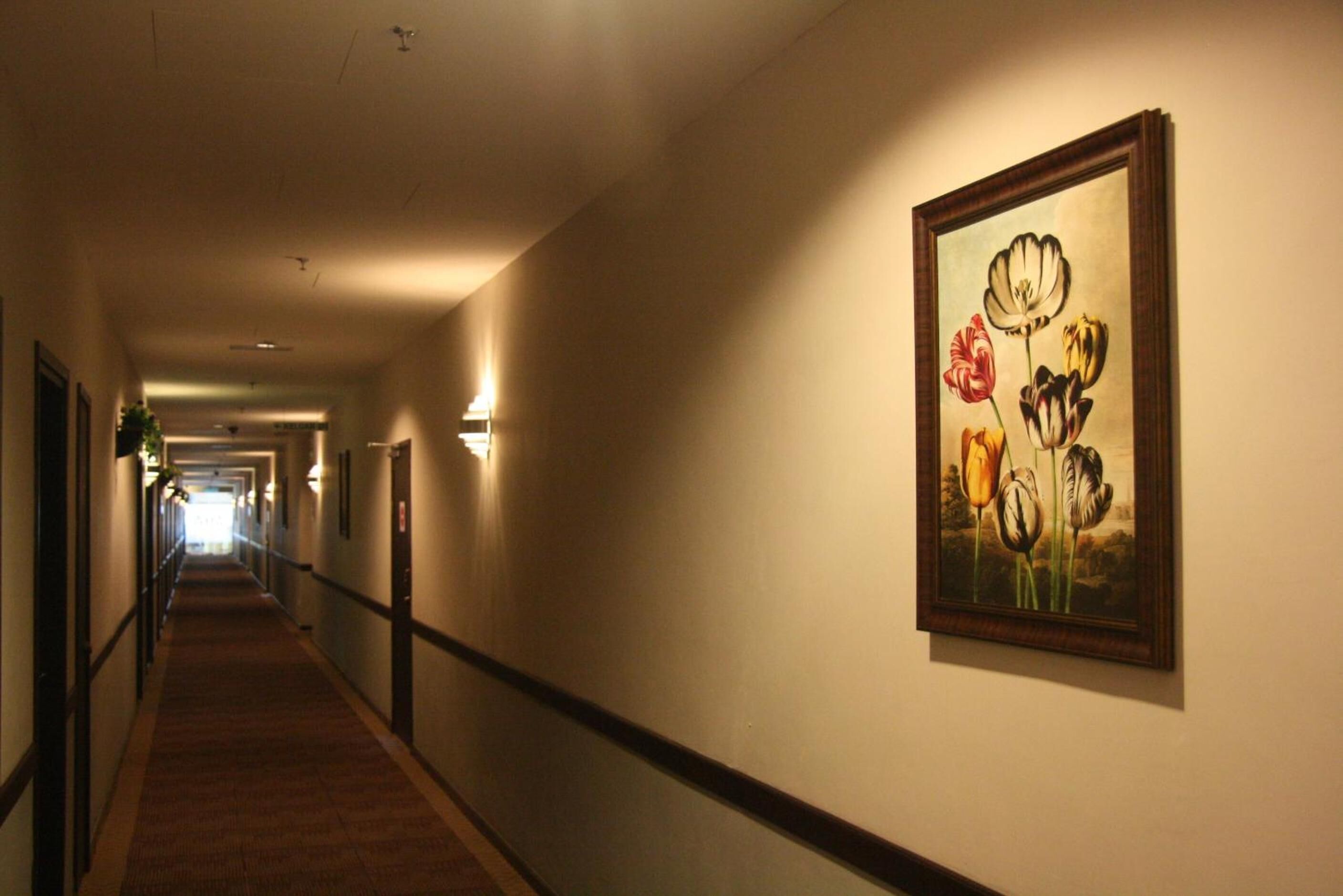 A hotel corridor with a carpeted floor, a painting of flowers on the wall, and a light fixture.