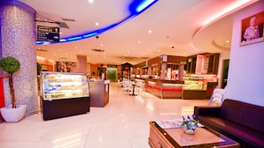 Un hall d’hôtel comprenant un comptoir d’accueil, un espace détente avec un canapé et une table basse, un présentoir avec des produits alimentaires, et des panneaux indicateurs pour les différents services, comme les toilettes et les chambres.