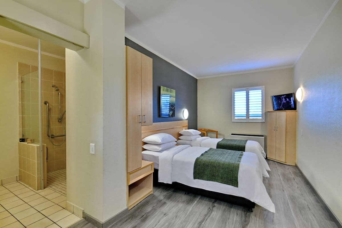 Ein Hotelzimmer mit zwei Betten, einer Duschkabine, einem Fernseher und einem Fenster mit Jalousien.