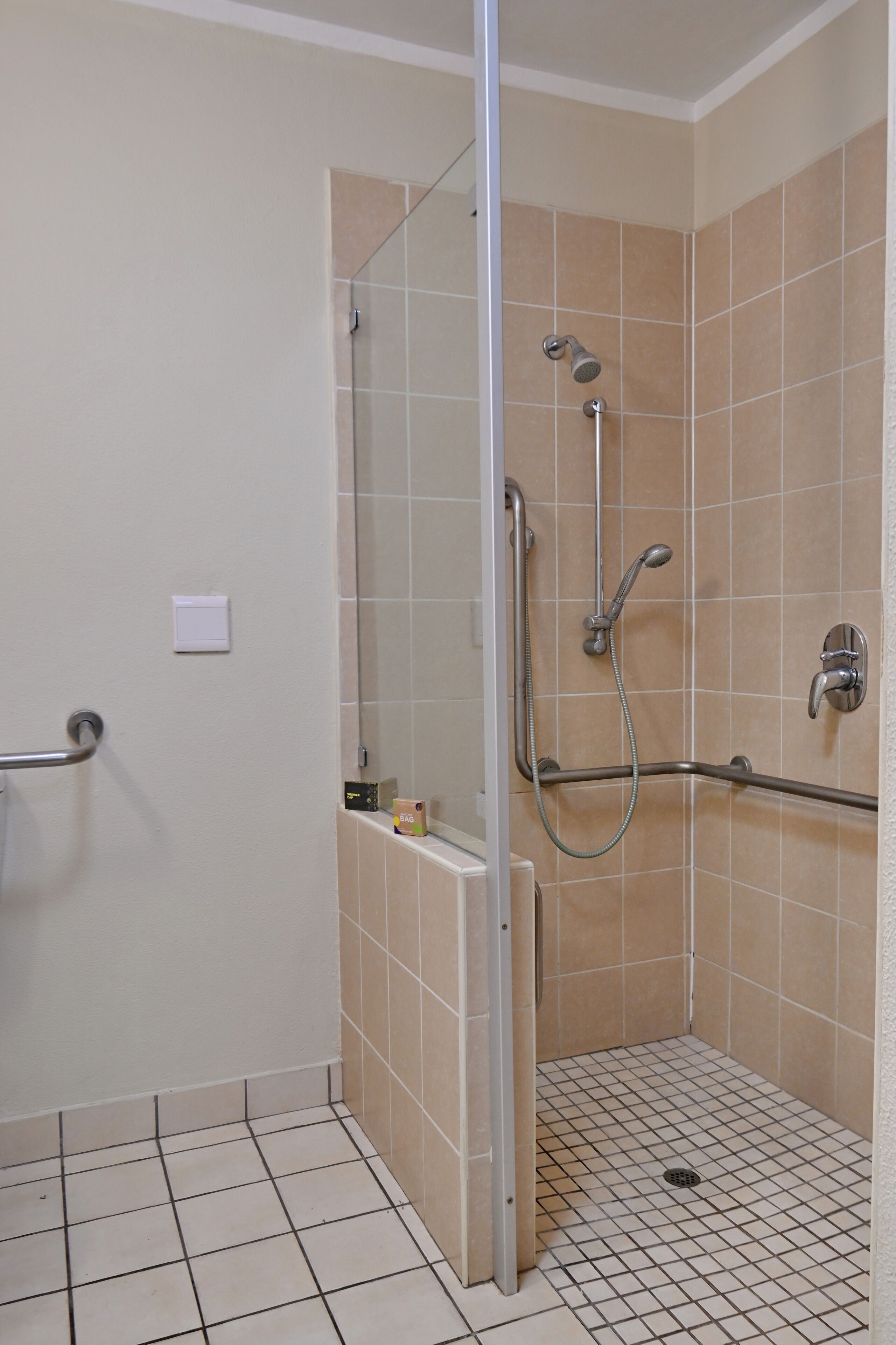 Un petit espace de douche carrelé, doté d’une porte en verre, d’une barre d’appui et d’un pommeau de douche.