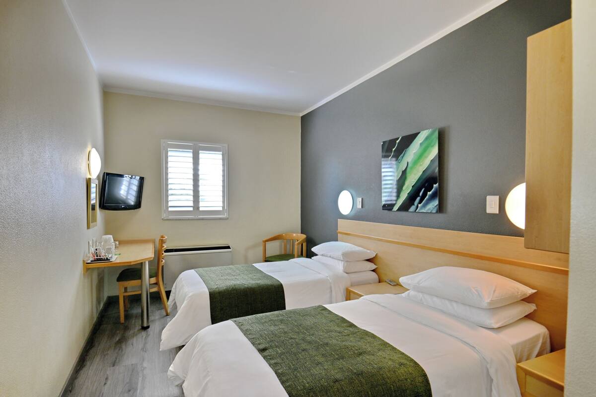 Ein Hotelzimmer mit zwei Betten, einem Schreibtisch, einem Stuhl, einem Fernseher und einem Fenster mit Jalousien.
