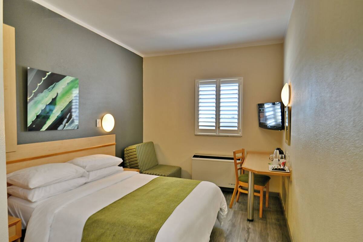 Ein Hotelzimmer mit Bett, Schreibtisch, Stuhl, Fernseher und einem Fenster mit Jalousien.