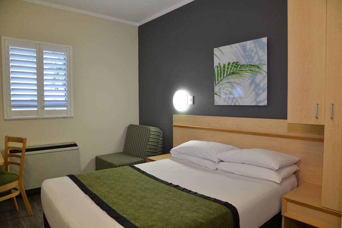 Ein Hotelzimmer mit einem Bett, einem Sessel, einem Schreibtisch, einer Heizung, einer Wandleuchte und einem Pflanzenbild.