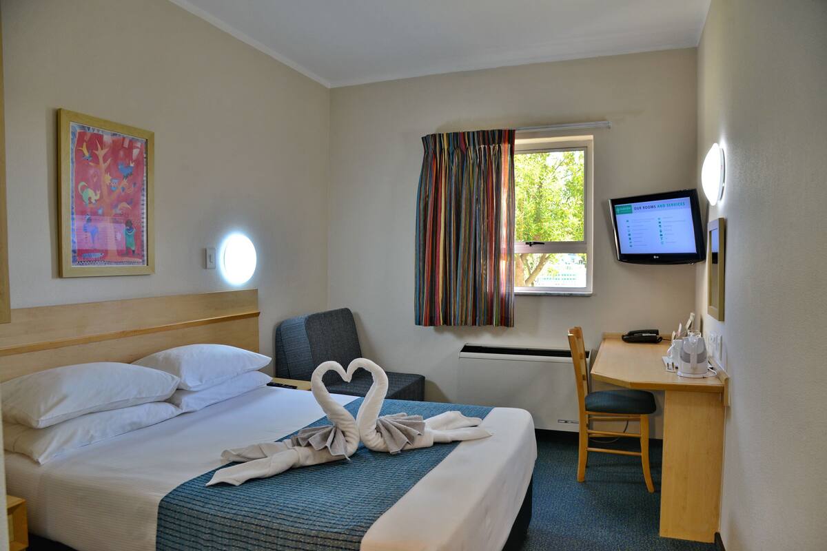 Ein Hotelzimmer mit Bett, Schreibtisch, Stuhl, Fernseher und einem fensterbepflanzten Fenster.