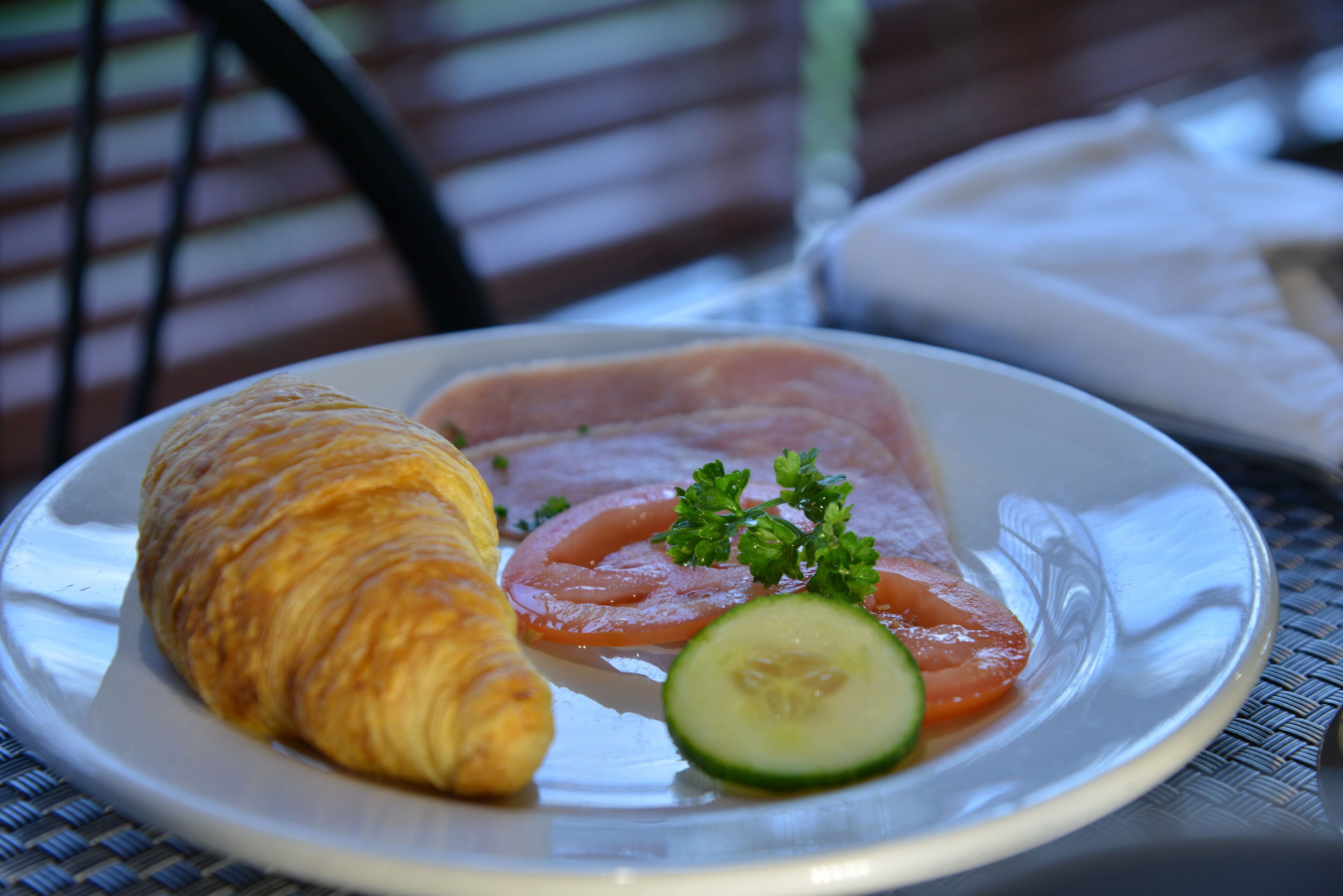 A plate with a croissant, sliced tomatoes, cucumber, and parsley.