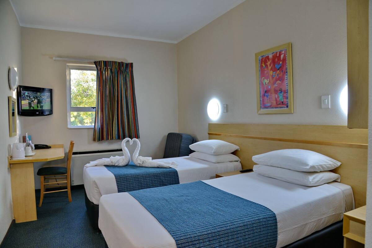 Ein Hotelzimmer mit zwei Betten, einem Schreibtisch, einem Stuhl, einem Fernseher und einem Fenster mit Vorhängen.