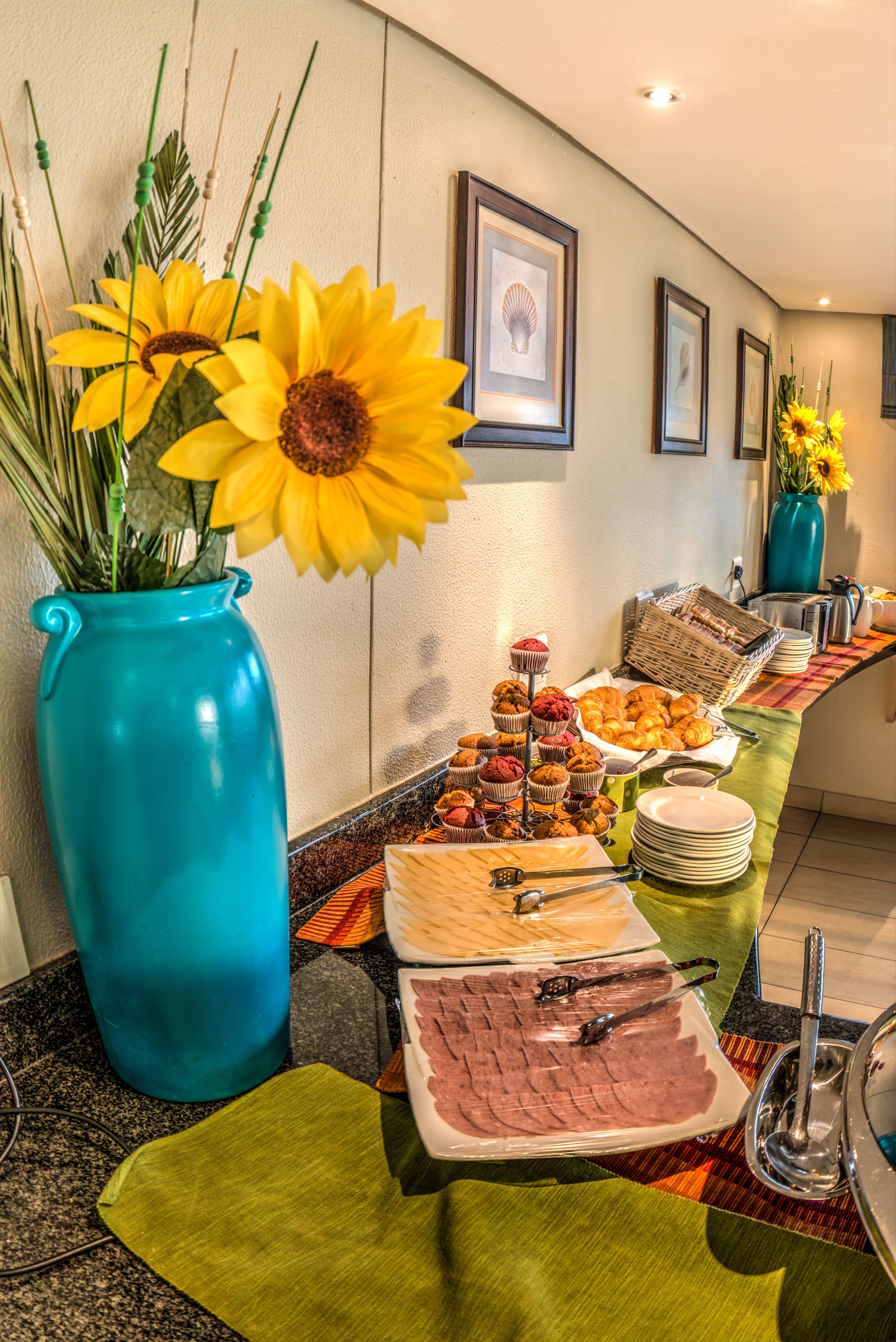 A buffet setup with various food items, a large blue vase with sunflowers, and framed artwork on the wall.