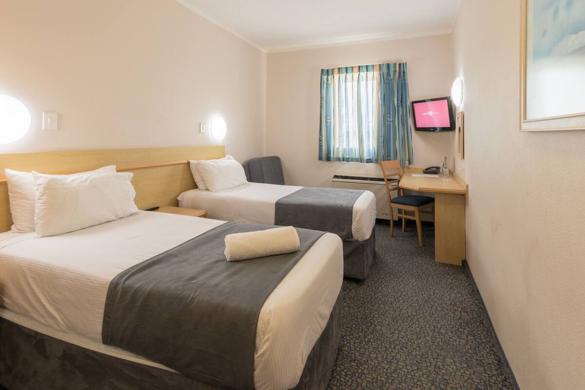 Ein Hotelzimmer mit zwei Betten, einem Schreibtisch, einem Stuhl, einem Fernseher und einem Vorhangfenster.