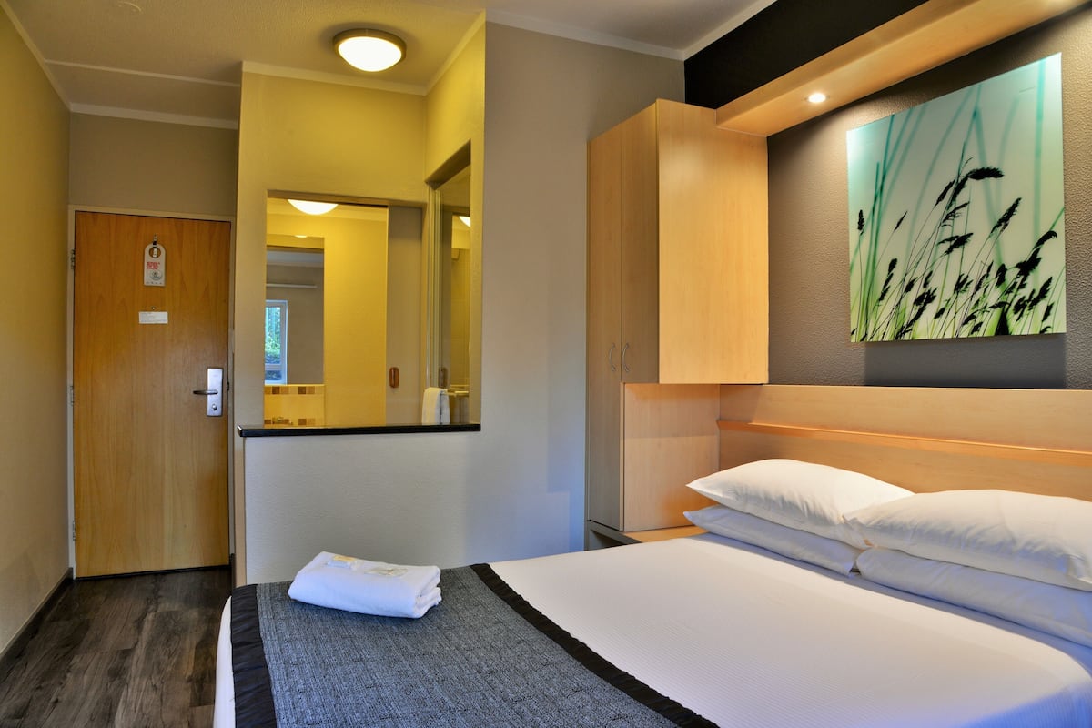 A hotel room with a bed, a wooden wardrobe, a mirror, and a wall-mounted artwork.