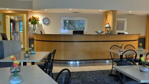 Un espace d’accueil d’hôtel avec un comptoir incurvé, une horloge et une œuvre d’art encadrée au-dessus. Plusieurs tables et chaises y sont disposées pour accueillir les clients.