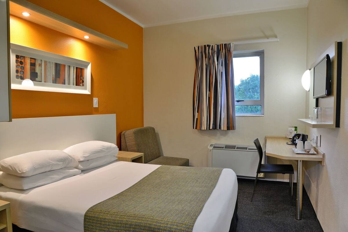A hotel room with a bed, a desk, a chair, a radiator, and a window with curtains.